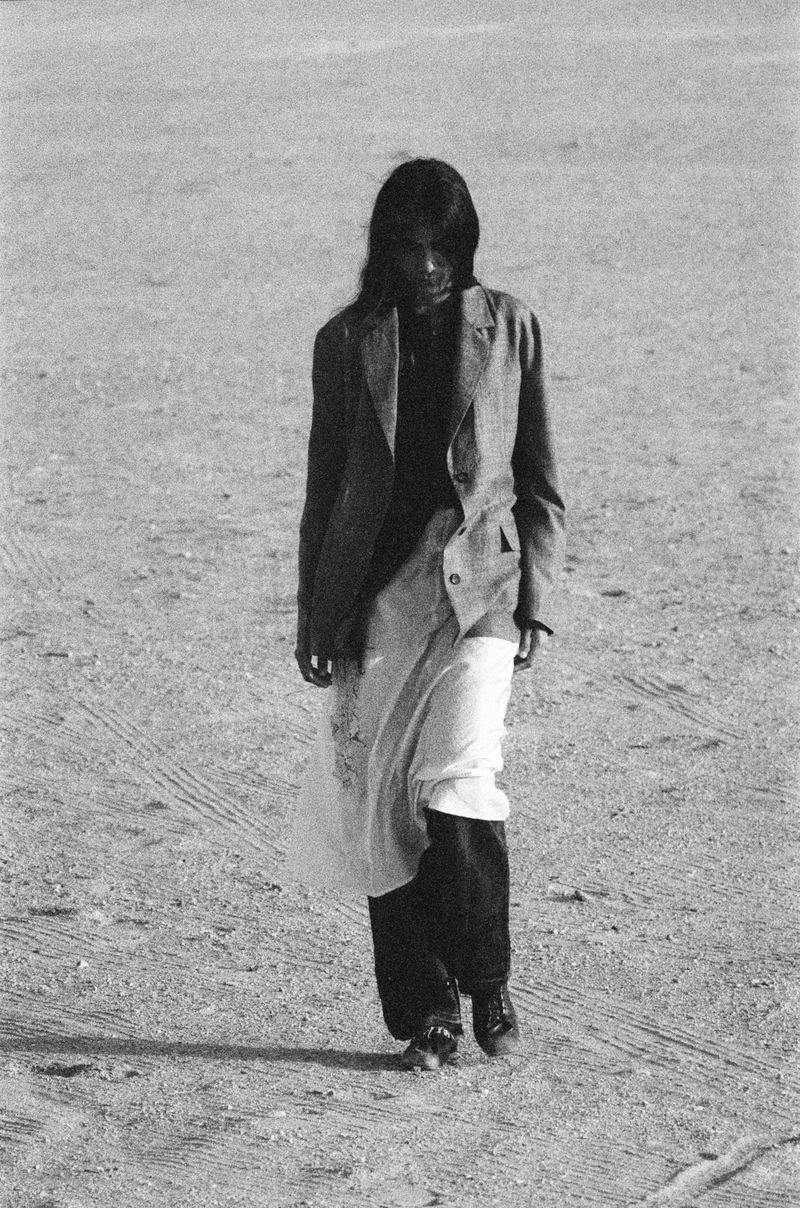 Model walking outdoors on sandy terrain wearing a tailored blazer and layered trousers for Kartik Research.
