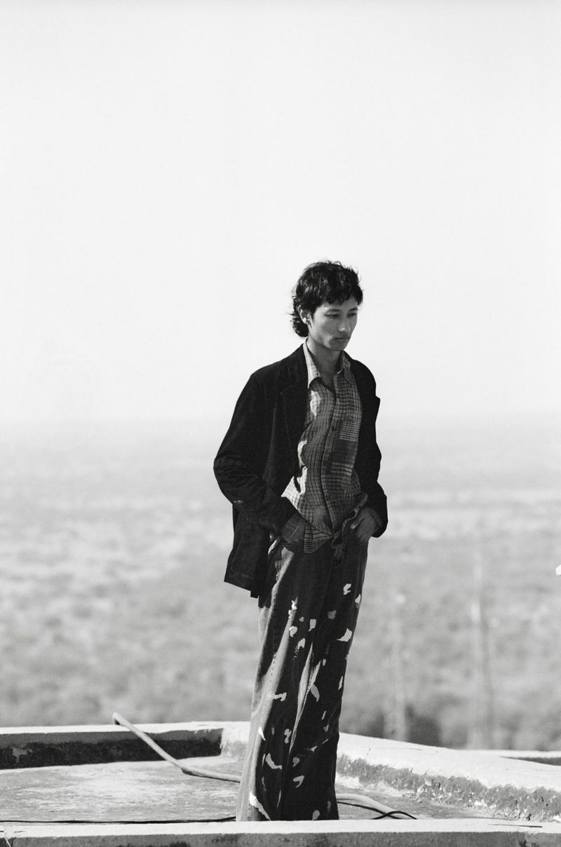 Man standing on a rooftop wearing a dark jacket and patterned trousers against an expansive landscape background