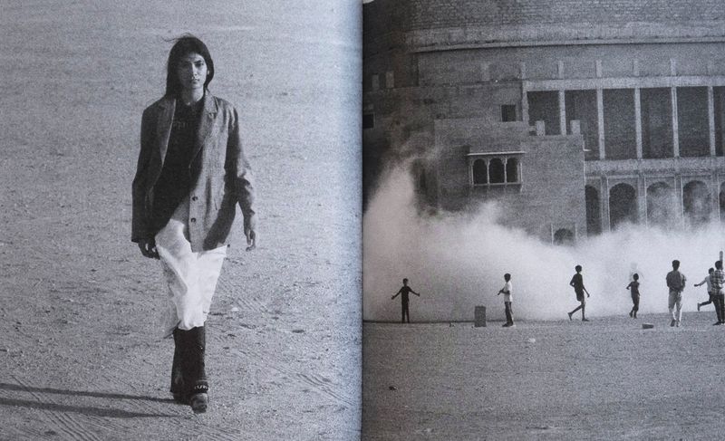 Model walking across a dusty ground past a large stone building with several figures in the background in a black and white campaign image