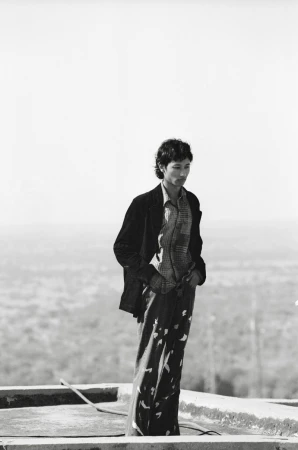 Man standing on a rooftop wearing a dark jacket and patterned trousers against an expansive landscape background