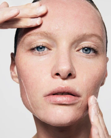 Close-up portrait of a fair-skinned adult female model with blue eyes, applying a translucent moisturizing treatment to her face with her fingers.