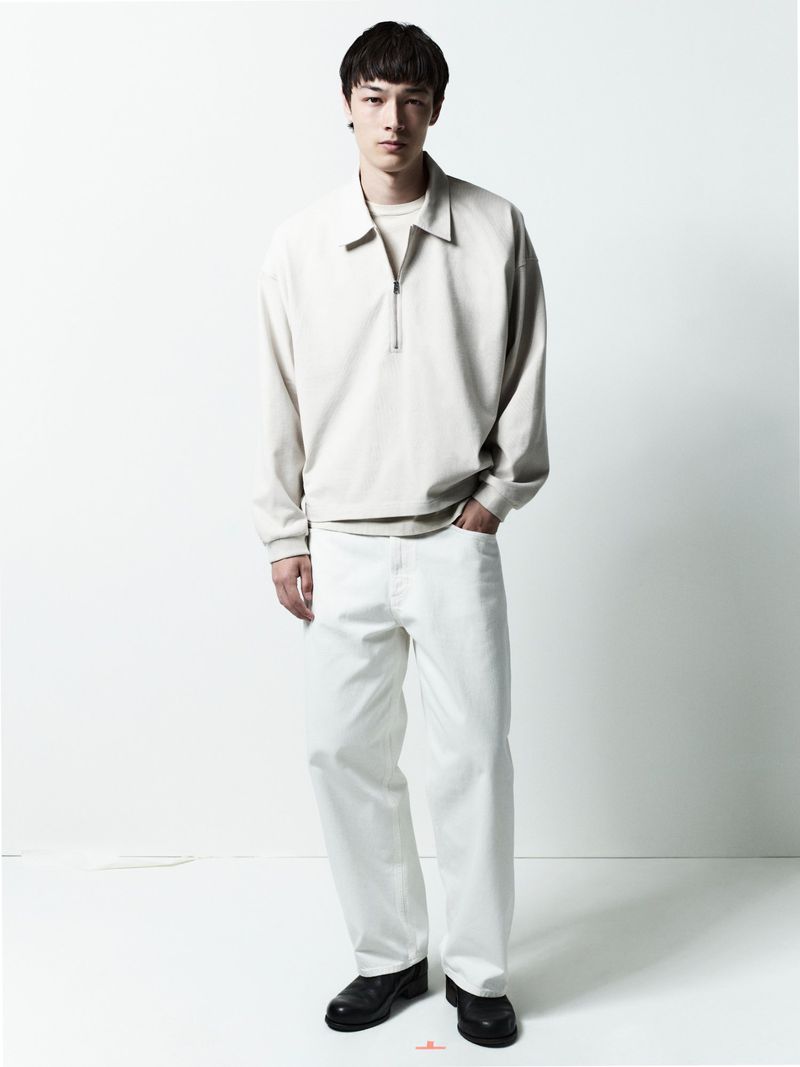 Male model wearing a cream-colored collared zip-up jacket and white jeans standing against a plain white studio backdrop.