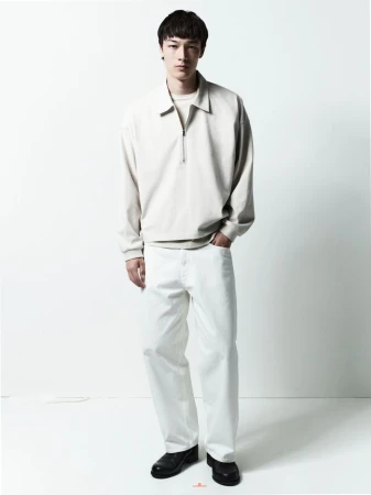 Male model wearing a cream-colored collared zip-up jacket and white jeans standing against a plain white studio backdrop.