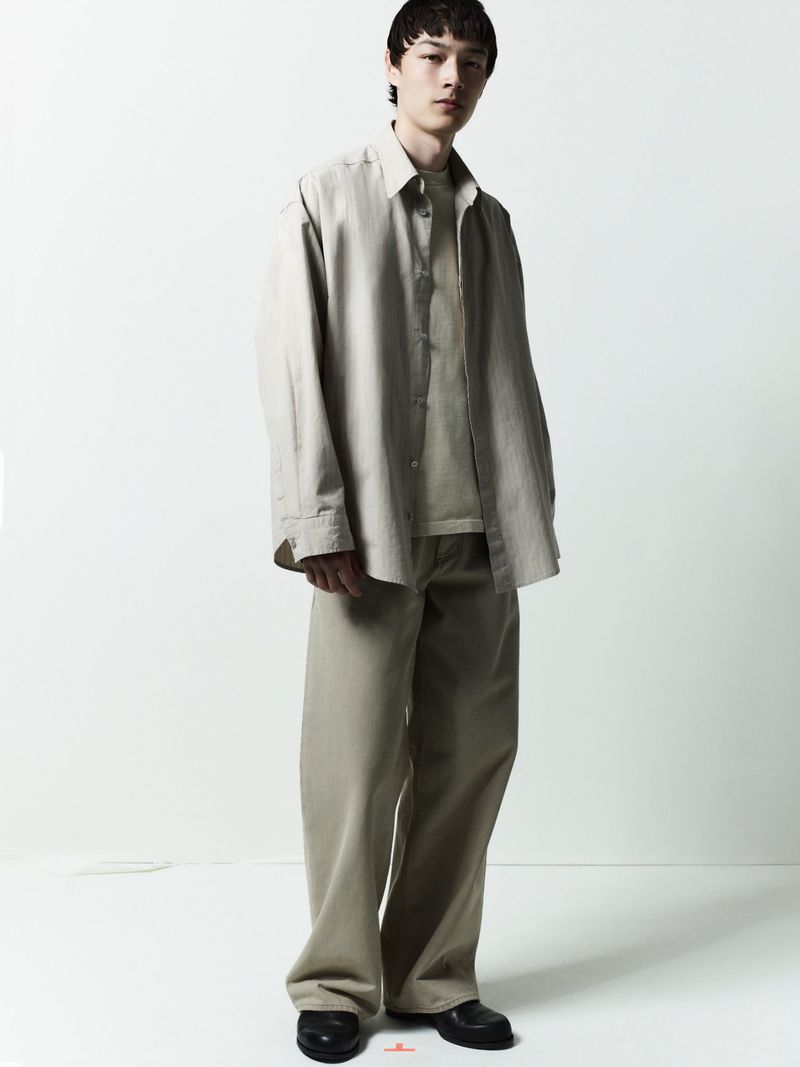 Full-length portrait of a male model wearing neutral-toned button-down shirt and relaxed trousers against a plain white background