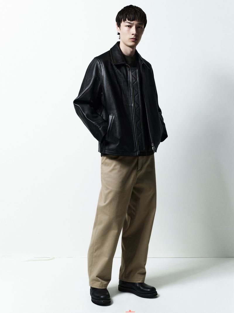 Model standing against a white seamless backdrop wearing a black leather jacket and tan trousers