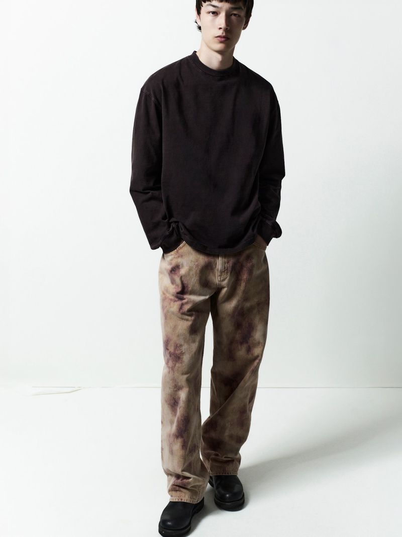 Male model in a dark long-sleeve shirt and patterned trousers standing against a white backdrop for Agolde Man.