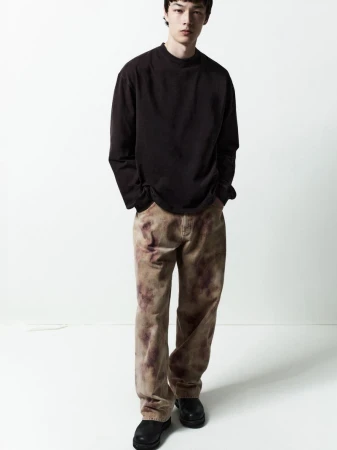 Male model in a dark long-sleeve shirt and patterned trousers standing against a white backdrop for Agolde Man.