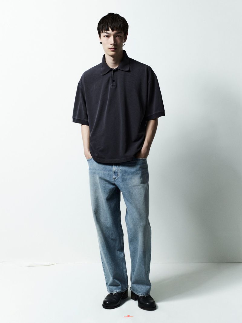 Model standing against a plain white background wearing a dark polo shirt and light blue denim jeans