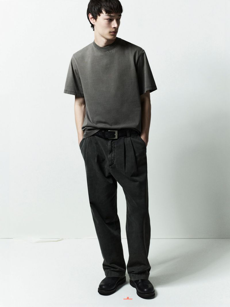 A male model wearing a grey t-shirt and dark trousers standing against a white studio background.