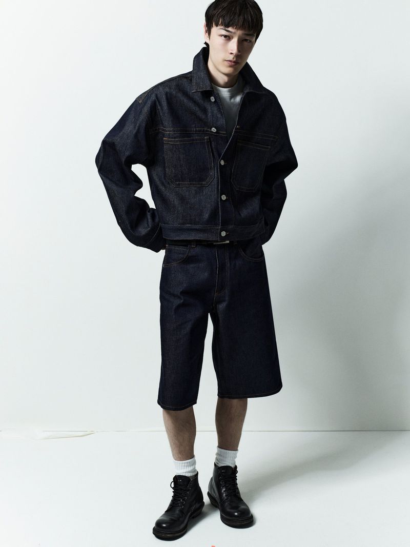 Full shot of a young adult Asian male in dark denim jacket and shorts, paired with socks and leather boots against a white backdrop.