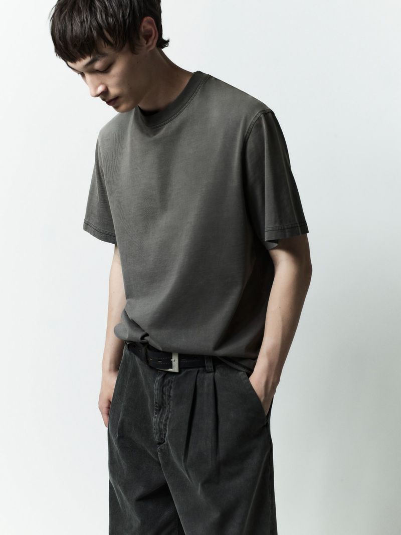 Young male model wearing a grey t-shirt and dark trousers against a white seamless background for AGOLDE Man