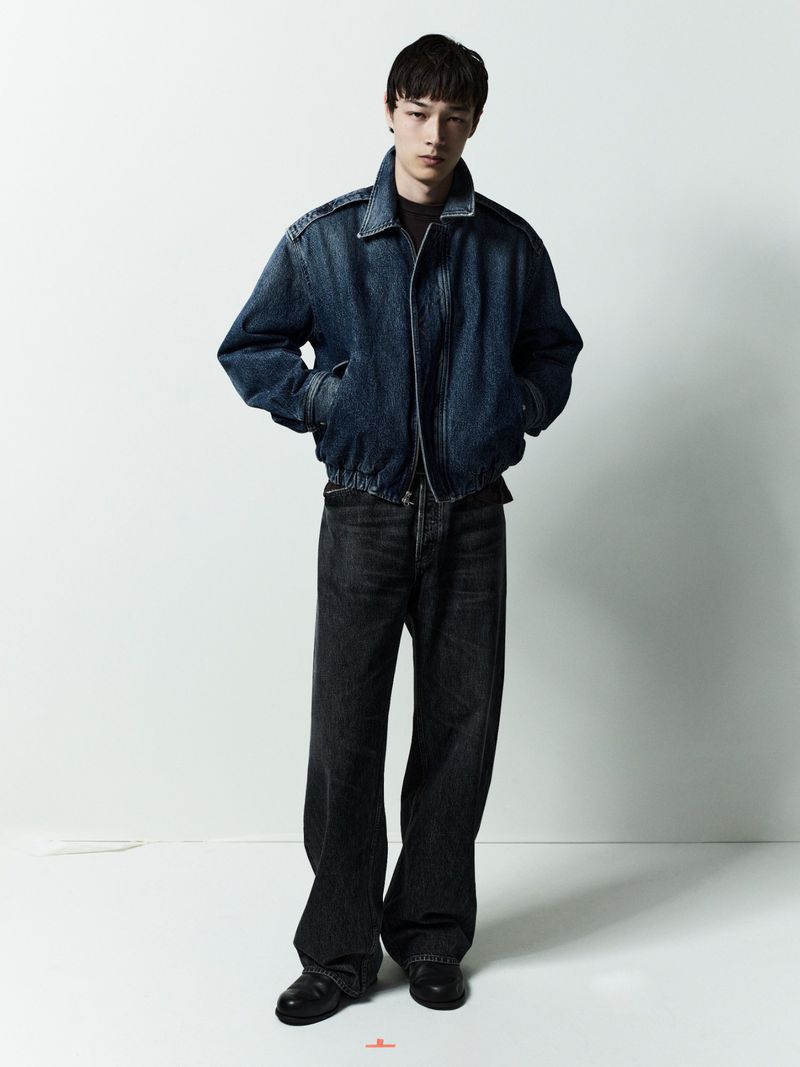 Model standing against a white studio background wearing a dark denim jacket and matching jeans.