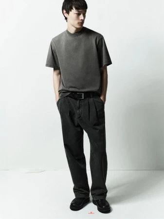 A male model wearing a grey t-shirt and dark trousers standing against a white studio background.