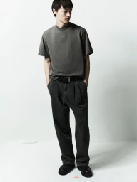 A male model wearing a grey t-shirt and dark trousers standing against a white studio background.