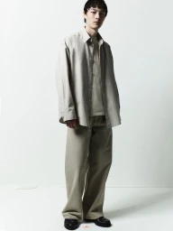 Full-length portrait of a male model wearing neutral-toned button-down shirt and relaxed trousers against a plain white background