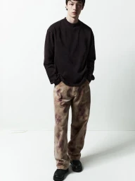 Male model in a dark long-sleeve shirt and patterned trousers standing against a white backdrop for Agolde Man.
