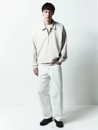 Male model wearing a cream-colored collared zip-up jacket and white jeans standing against a plain white studio backdrop.