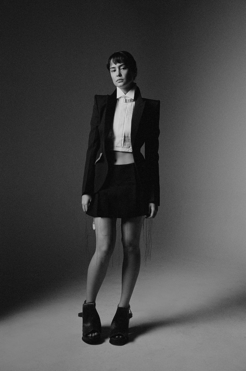 A young woman poses in a black blazer, white crop top, and black miniskirt in a studio setting, with a grayscale color palette.