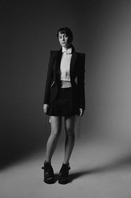 A young woman poses in a black blazer, white crop top, and black miniskirt in a studio setting, with a grayscale color palette.