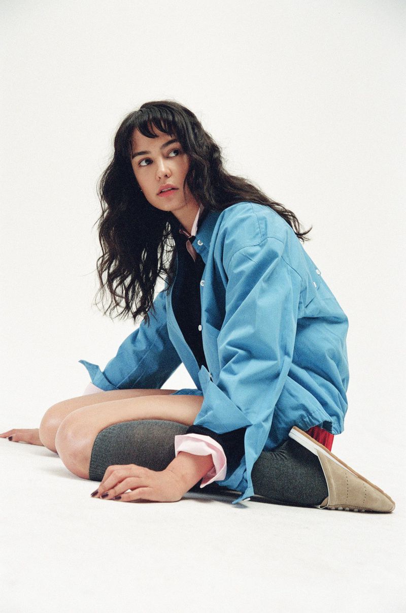 Brunette woman in a light blue jacket poses on a white background, looking slightly to her left.  She is sitting with knees bent.