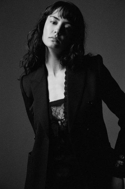 Monochromatic portrait of a woman with bangs, wearing a dark blazer and lace top, gazing at the camera against a neutral background.