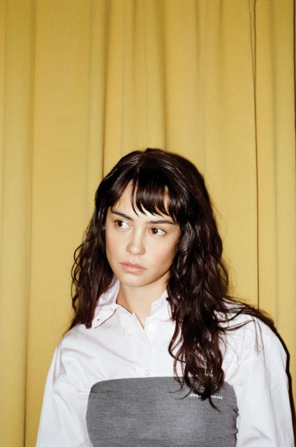 Portrait of a young adult woman with long brown hair wearing a white shirt and gray top against a yellow curtain backdrop.