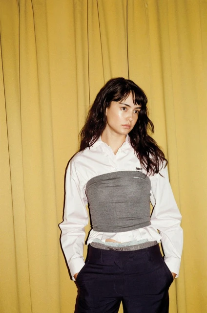A woman in a gray bandeau top and dark pants poses against a yellow curtain, presenting a fashionable and visually striking image.
