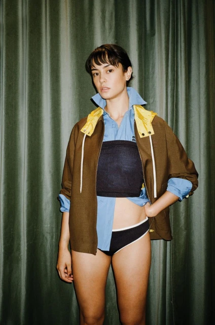 Young adult woman poses in a blue shirt and black briefs with a jacket over top in a studio with green curtains.