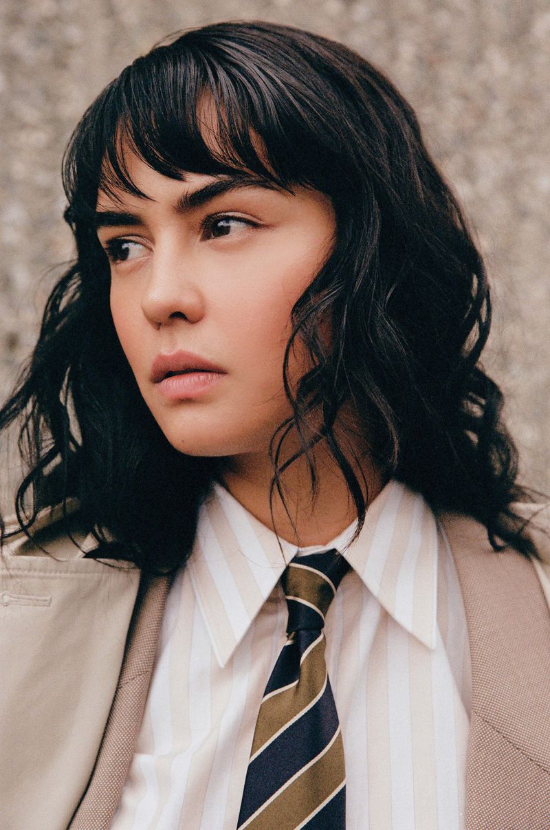 Portrait of a woman with dark hair and bangs wearing a striped shirt, tie, and trench coat against a textured grey wall, looking left.