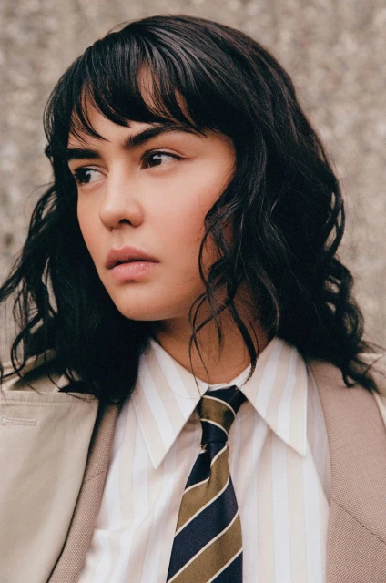 Portrait of a woman with dark hair and bangs wearing a striped shirt, tie, and trench coat against a textured grey wall, looking left.