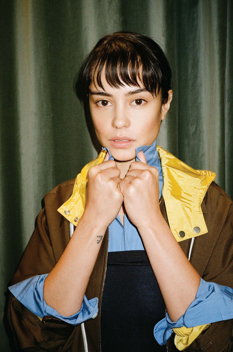 Portrait of a young woman with bangs wearing a brown and yellow jacket against a green curtain. She is looking directly at the camera.