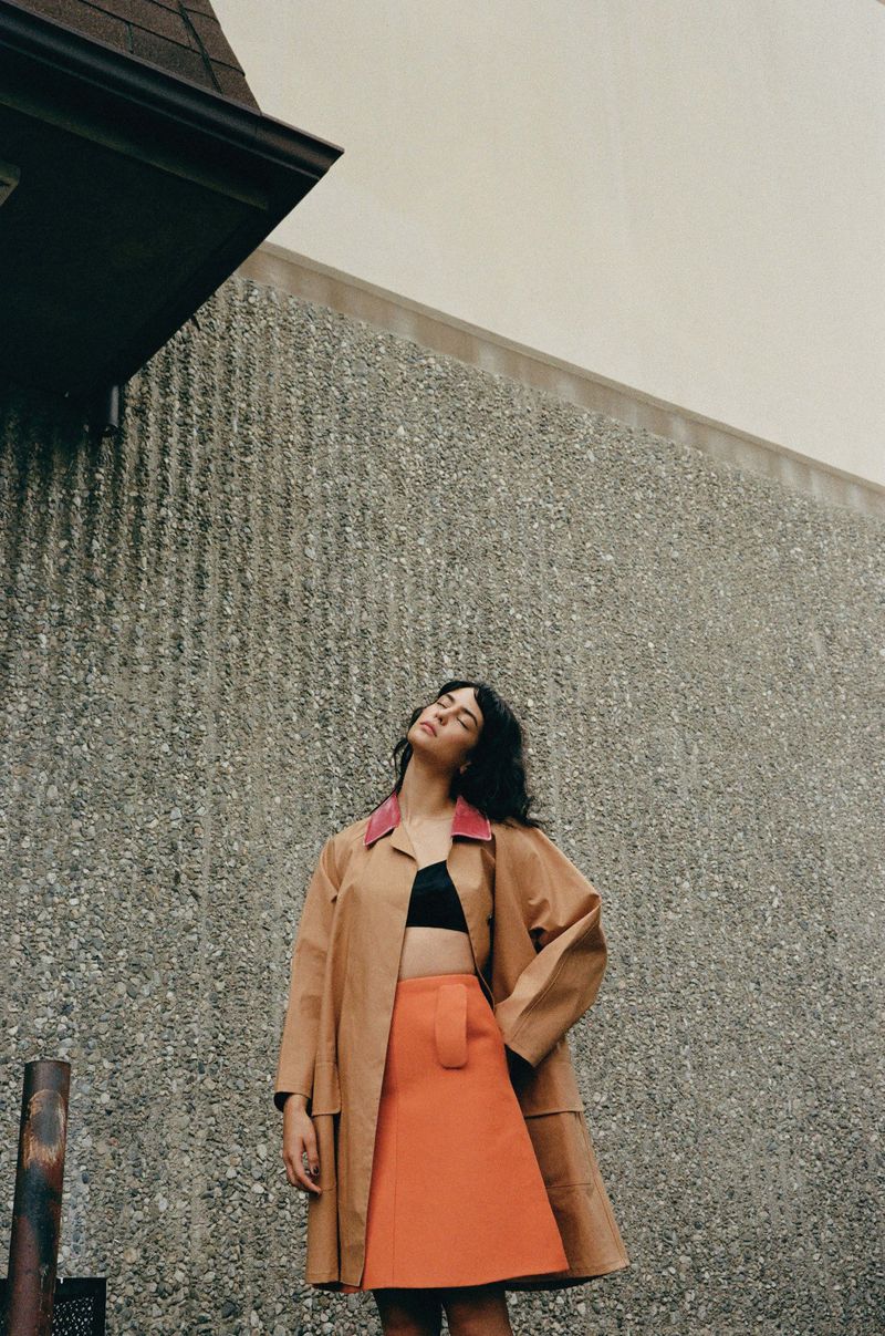 Woman in a tan coat and orange skirt poses against a gray wall with her head tilted back and eyes closed in an outdoor setting.