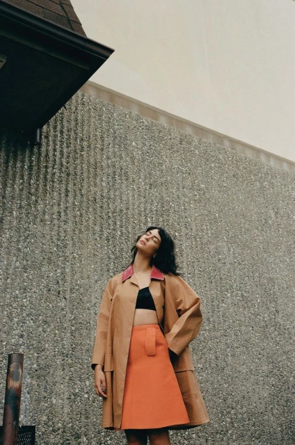 Woman in a tan coat and orange skirt poses against a gray wall with her head tilted back and eyes closed in an outdoor setting.