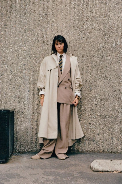 A woman poses in a tan trench coat over a brown double-breasted suit against a mottled concrete wall in a full outdoor shot.