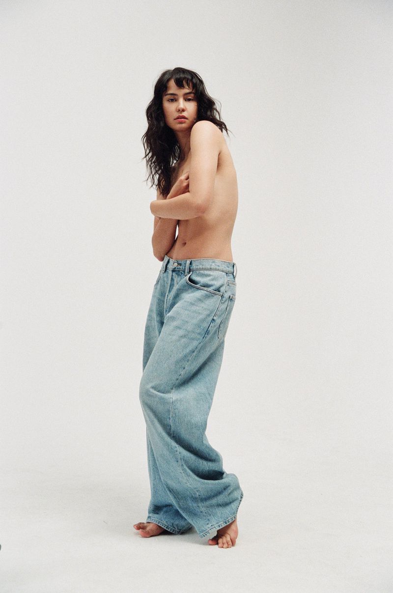 A young woman poses in light blue, wide-leg denim jeans against a white backdrop, showcasing casual fashion in a studio setting.