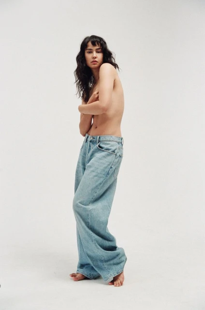 A young woman poses in light blue, wide-leg denim jeans against a white backdrop, showcasing casual fashion in a studio setting.