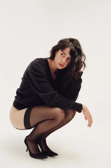 Portrait of a brunette woman crouching indoors, wearing a black sweater, sheer black stockings, and black high heels.