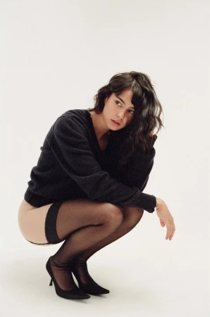 Portrait of a brunette woman crouching indoors, wearing a black sweater, sheer black stockings, and black high heels.