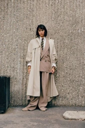 Model standing against a textured concrete wall wearing an oversized beige trench coat over a suit and tie