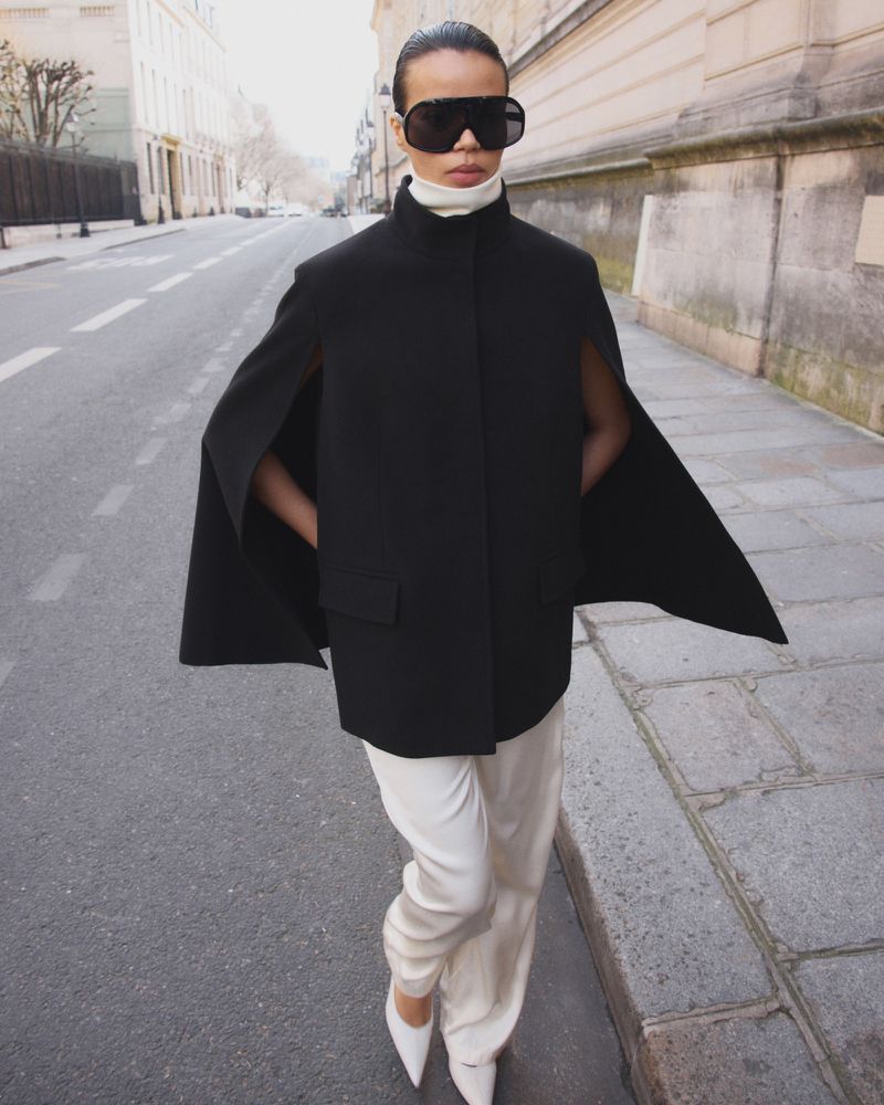 Model wearing a black cape coat, white trousers, and oversized sunglasses walking on a city street.