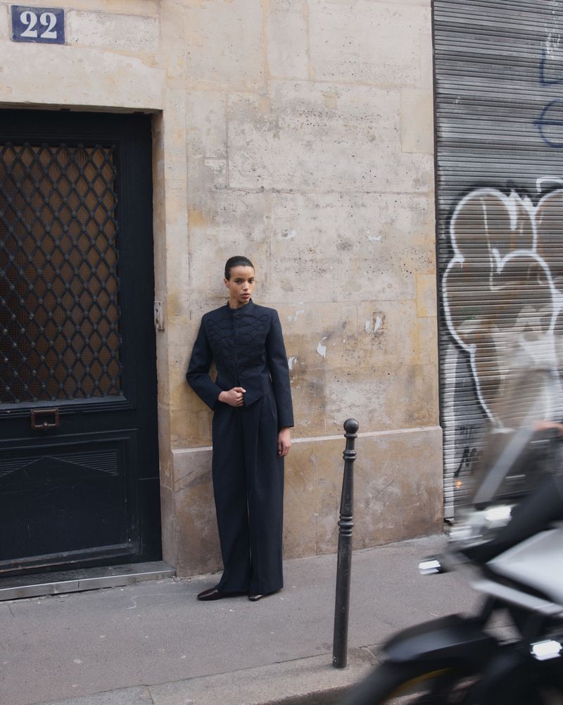 Model standing on a city sidewalk leaning against a stone building wall next to a dark door and graffiti-covered shutter