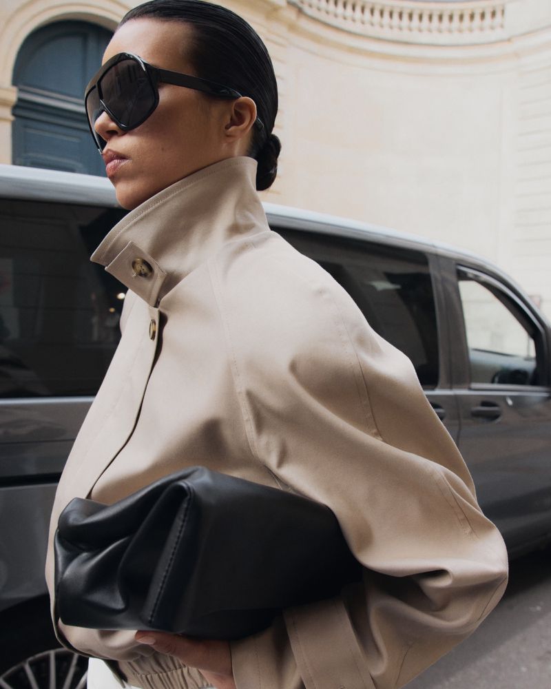 Stylish woman in beige jacket, sunglasses, holds black handbag, standing in city street with car and architectural building visible.