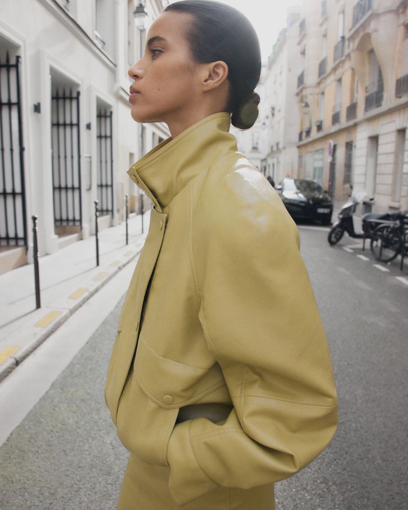 Profile view of a model in a pale yellow jacket standing on an urban street.