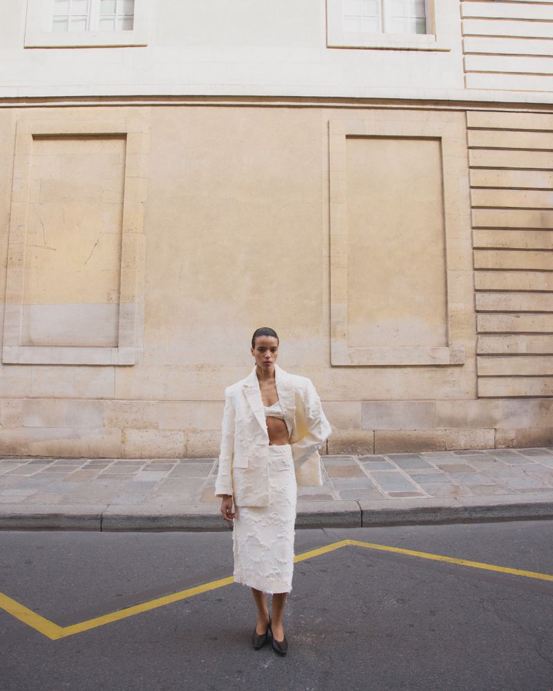 Model standing on a paved street wearing a white textured blazer and skirt set against a tall classical stone building.