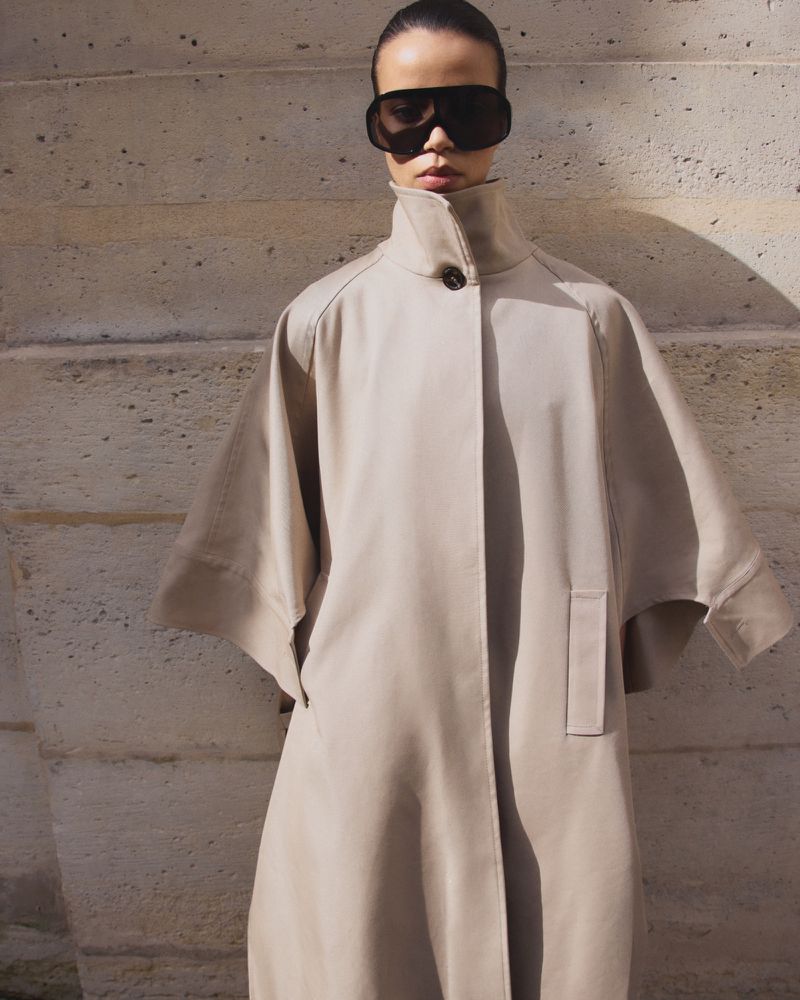 Model wearing dark oversized sunglasses and a beige cape coat standing in front of stone architecture.