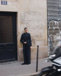 Model standing on a city sidewalk leaning against a stone building wall next to a dark door and graffiti-covered shutter