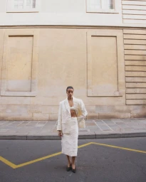 Model standing on a paved street wearing a white textured blazer and skirt set against a tall classical stone building.