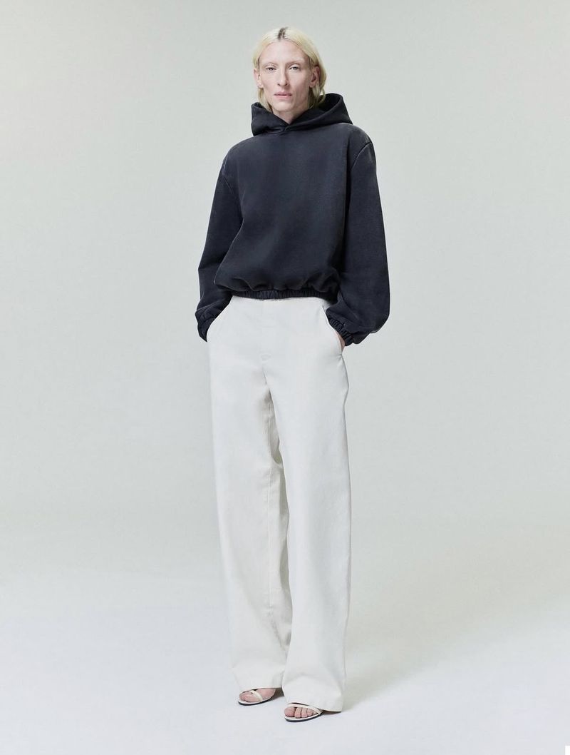 Model wearing a dark hoodie and white trousers standing against a seamless light grey studio background.