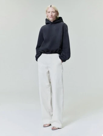 Model wearing a dark hoodie and white trousers standing against a seamless light grey studio background.
