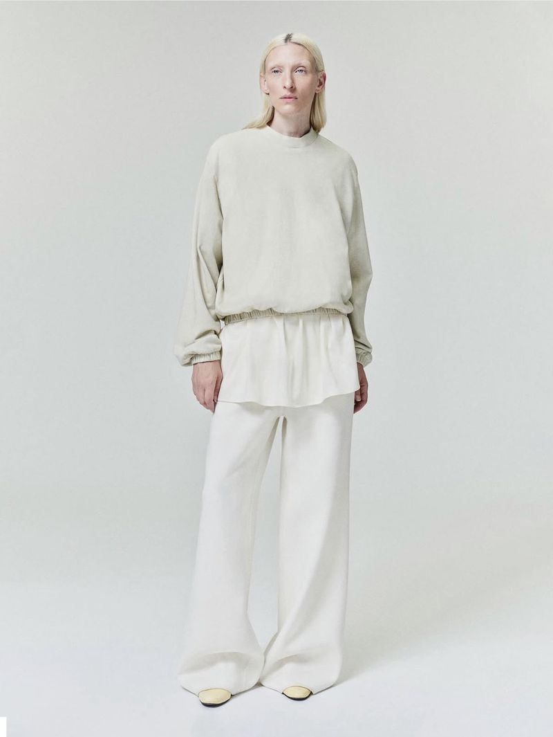 Model wearing neutral toned layered sweatshirt and loose trousers standing against a pale grey seamless studio backdrop